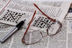 A newspaper with a crossword puzzle, a pen, and eyeglasses on top of the newspaper used as a distraction to reduce boredom during covid 19