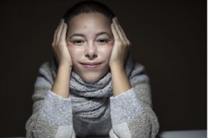 A teenage girl with a dark background sitting along with her hands to her cheeks with a look of boredom on her face