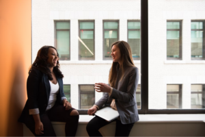 two women talking smiling sitting on window sill