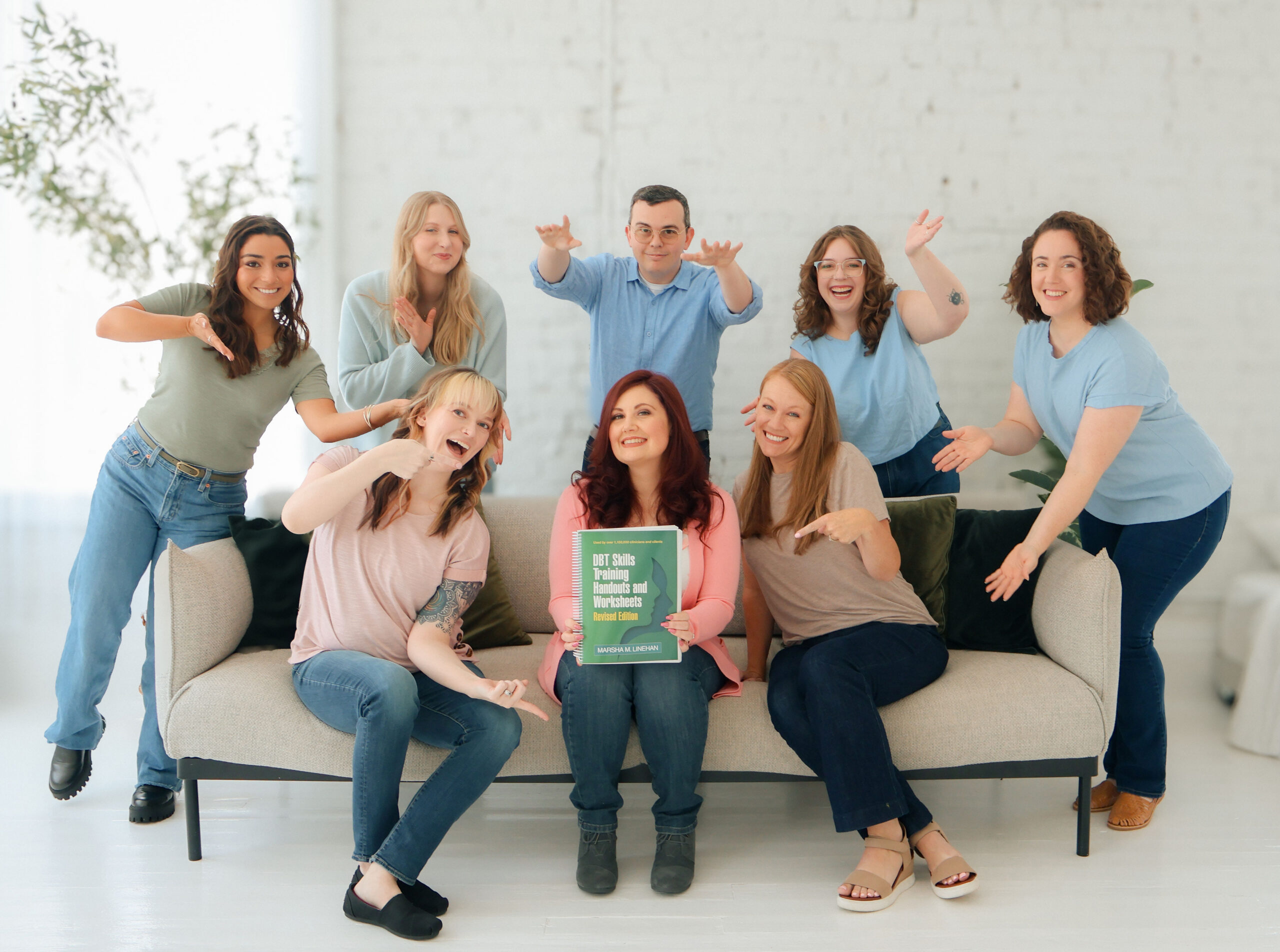 Cincinnati Center for DBT DBT clinicians posing as a group. Eight DBT therapists sit on a couch pointing at the DBT Skills Training Handouts and Worksheets book.